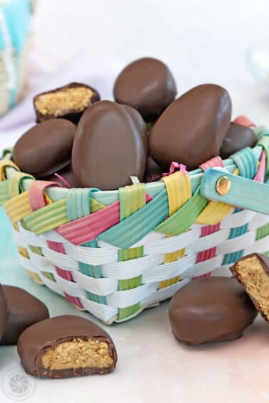 Peanut butter Easter eggs in and around a colorful Easter basket with a few eggs cut in half to show center filling.